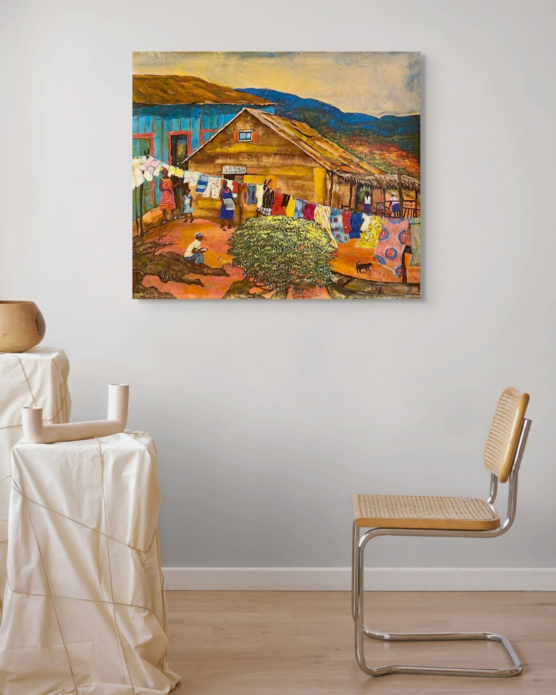 Painting of a rustic scene with a barn and people hanging laundry, displayed on a wall in a room with a chair and table.