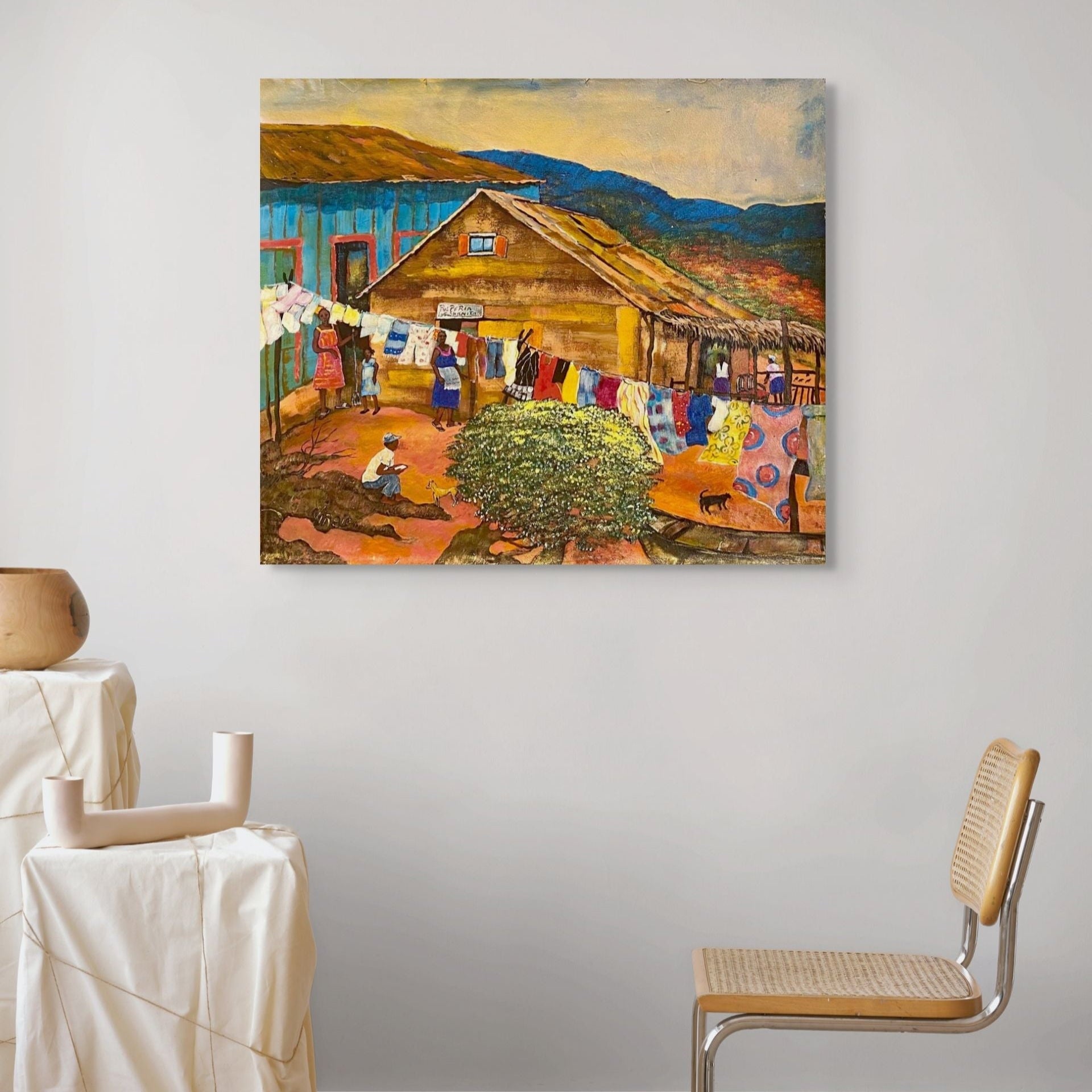 Painting of a rustic scene with a barn and people hanging laundry, displayed on a wall in a room with a chair and table.