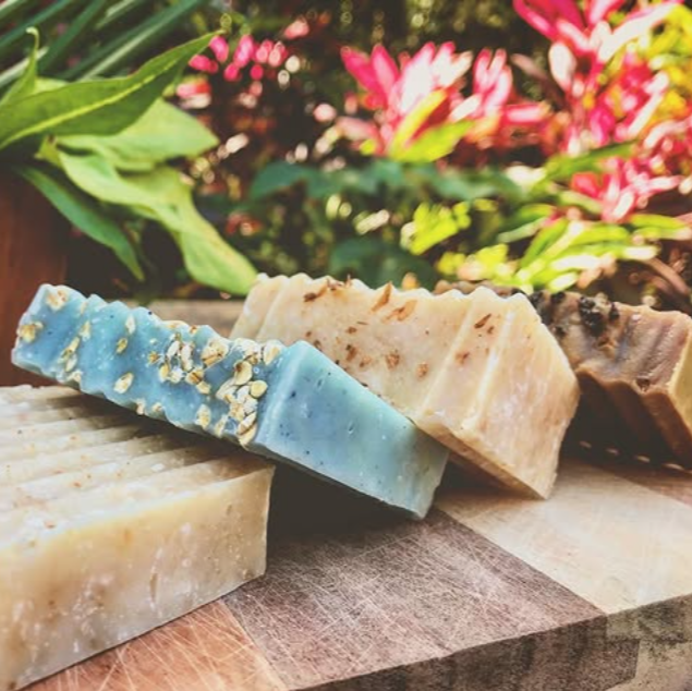 Three bars of soap, one blue with specks and two beige, on a wooden surface with a floral background.