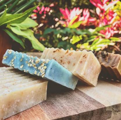 Three bars of soap, one blue with specks and two beige, on a wooden surface with a floral background.