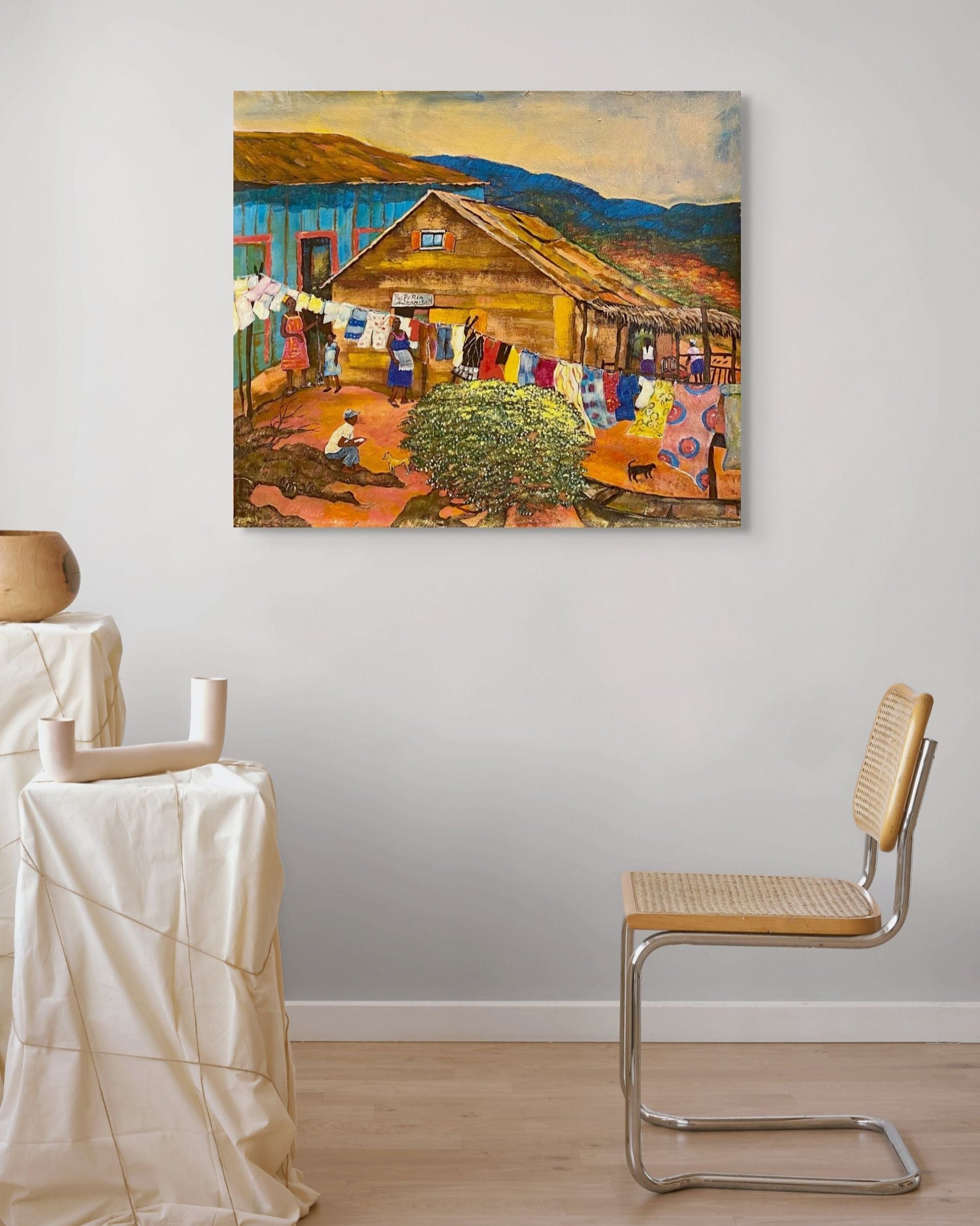 Painting of a rustic scene with a barn and people hanging laundry, displayed on a wall in a room with a chair and table.