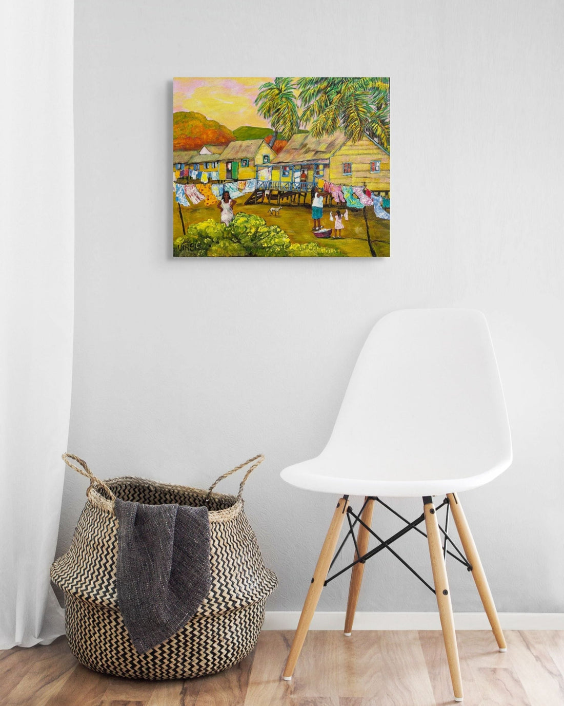 Colorful painting of a village scene on a white wall above a woven basket and white chair.